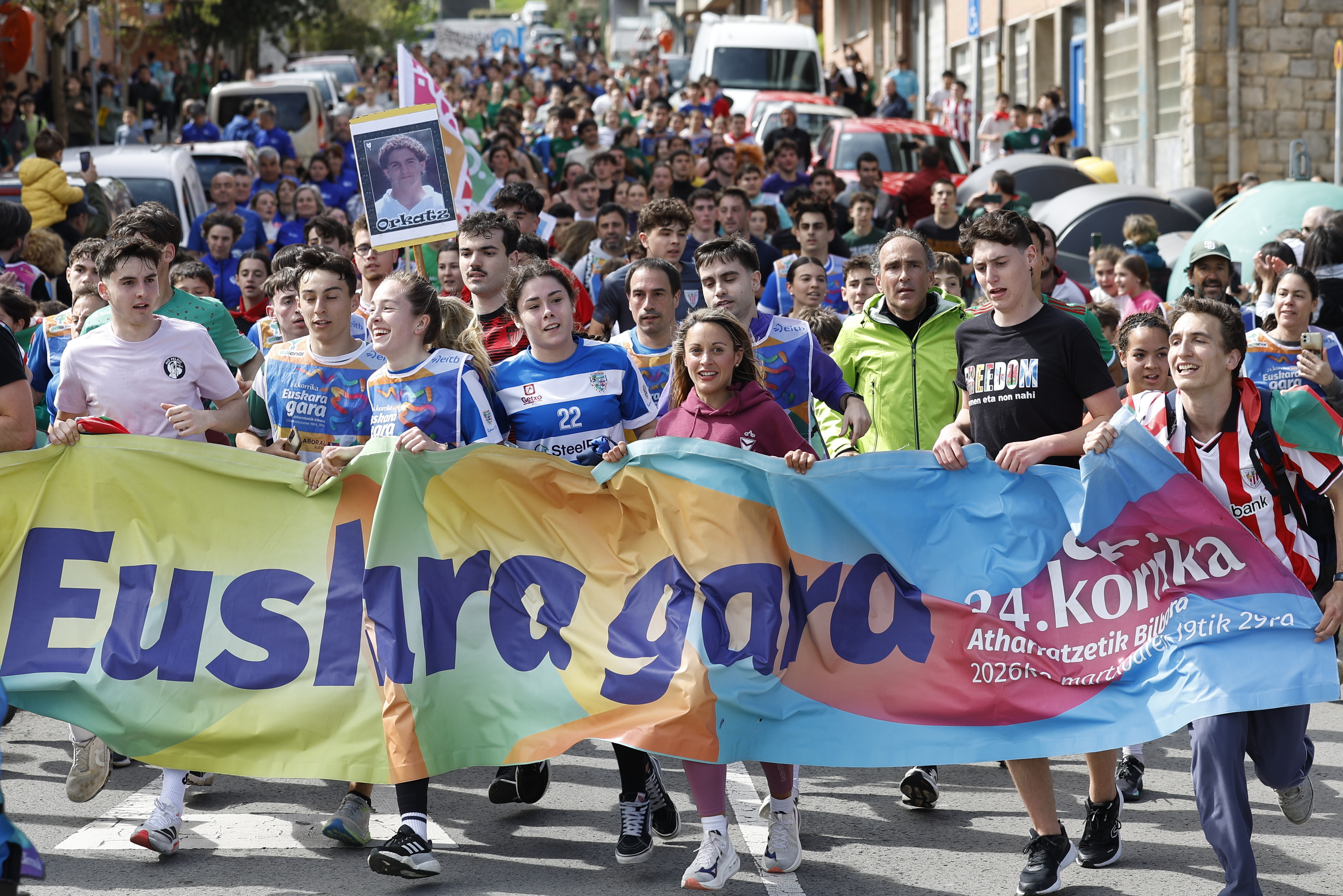 Varios jóvenes llevan la pancarta de la Korrika por delante de una fotografía de Orkatz Gallastegi