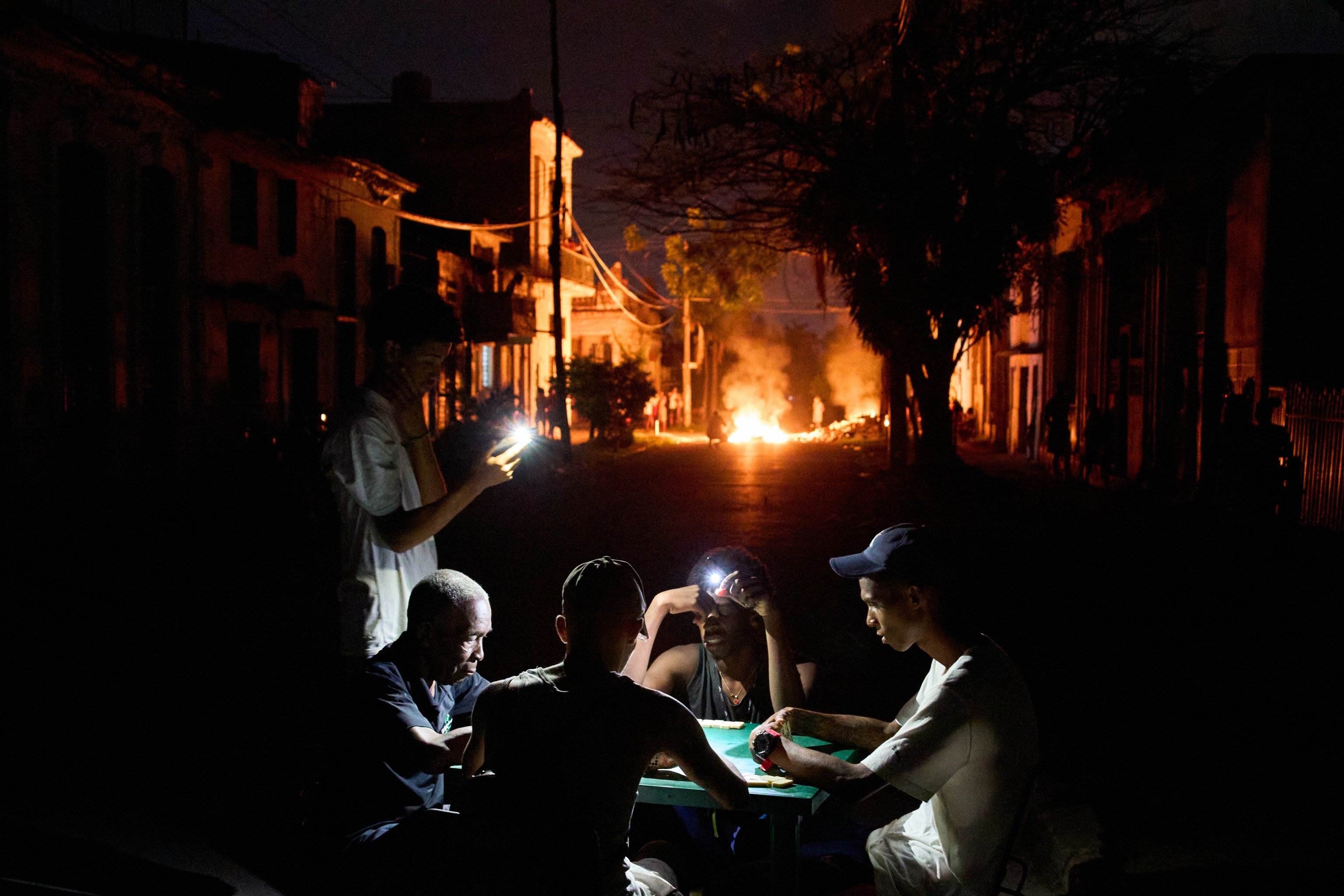 Varias personas se iluminan con sus teléfonos mientras juegan al dominó, al tiempo que arde en una calle de La Habana.