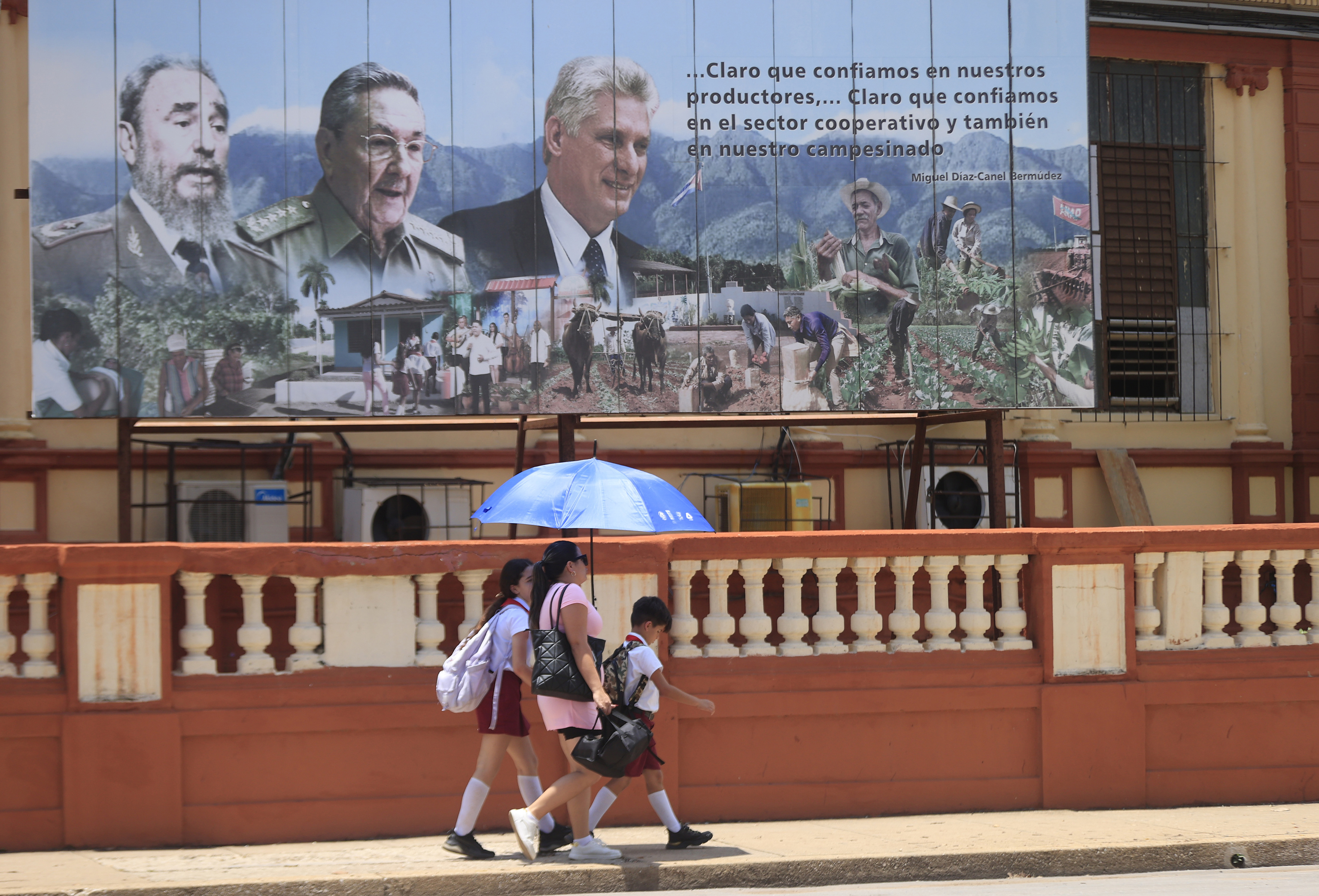 na mujer camina con dos niños en La Habana frente a un mural en el que están pintados Fidel y Raúl Castro, y Miguel Díaz-Canel.